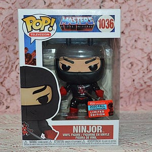 May include: A Ninjor vinyl figure from the Masters of the Universe series, packaged in a box. The figure is black and red, with a ninja mask. The box includes the Pop! Television logo and a 2020 Fall Convention Limited Edition sticker.