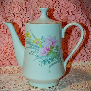 May include: A white porcelain coffee pot with a lid, featuring a floral design in pink, yellow, and blue. The pot has a curved handle and a gold trim around the base and the rim of the lid. The background is a pink lace fabric.