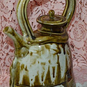 May include: A handcrafted ceramic teapot with a twisted handle and a lid. The teapot is primarily brown with white accents and a unique, textured design. The spout and handle are a lighter shade of brown.