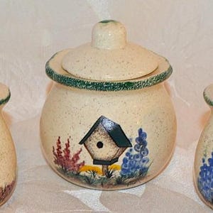 May include: A ceramic kitchen set featuring a speckled beige finish. The set includes two salt and pepper shakers and a lidded jar. Each piece is decorated with a birdhouse and floral design in shades of blue, red, and yellow.