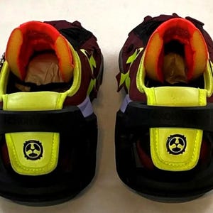 May include: Pair of futuristic sneakers with a black, yellow, and burgundy color scheme. The shoes feature a unique design with a black sole, yellow accents, and a burgundy upper. The tongue and inner lining are bright orange. The heels have a black strap with a yellow emblem.