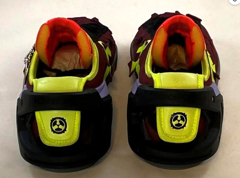 May include: Pair of futuristic sneakers with a black, yellow, and burgundy color scheme. The shoes feature a unique design with a black sole, yellow accents, and a burgundy upper. The tongue and inner lining are bright orange. The heels have a black strap with a yellow emblem.