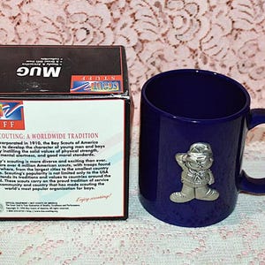 May include: A navy blue ceramic mug with a silver-colored Scout emblem. The mug is next to a box with the words "SCOUT STUFF" and "MUG" printed on it. The box has text about the Boy Scouts of America.