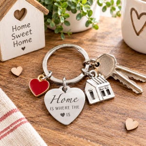 May include: A silver-toned metal keyring featuring a heart-shaped charm engraved with the text 'Home IS WHERE THE HEART IS', a small red enamel heart charm, a miniature house charm, and two silver keys attached to a circular split ring.