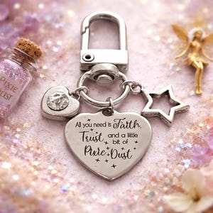 May include: A silver keyring with a heart pendant engraved with "All you need is Faith, Trust, and a little bit of Pixie Dust." The keyring also features a heart charm with a crystal and a star charm. A small bottle of pink glitter is visible.
