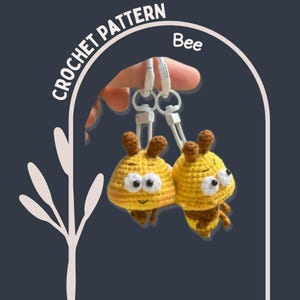 May include: Two yellow and brown crochet bee keychains with white metal clasps. The bees have smiling faces and are labeled "CROCHET PATTERN Bee". The keychains are handmade and feature a whimsical design.