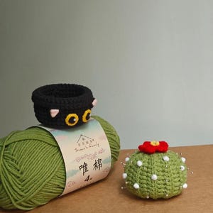 May include: A black crochet cat-shaped basket with yellow eyes and pink ears sits atop a green yarn ball. A green crochet cactus with a red flower and white dots is also present. The yarn label has Chinese text.