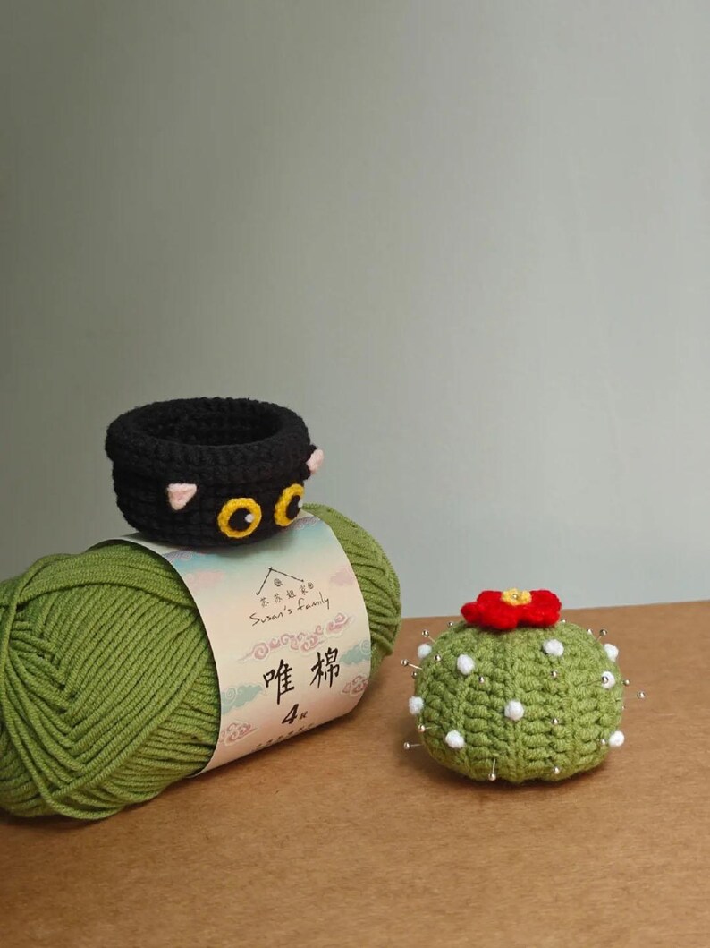 May include: A black crochet cat-shaped basket with yellow eyes and pink ears sits atop a green yarn ball. A green crochet cactus with a red flower and white dots is also present. The yarn label has Chinese text.