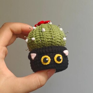 May include: A handmade crochet pincushion shaped like a cactus with a black cat base. The cactus is green with white accents and red flowers. The cat has yellow eyes and pink ears. Silver pins are inserted into the cactus.