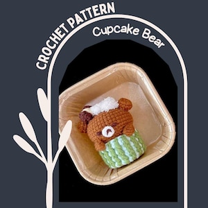 May include: A crocheted brown bear with a white and brown topping, nestled in a green and white striped cupcake liner. The bear is in a small, square, paper container. The words "CROCHET PATTERN" and "Cupcake Bear" are in the image.