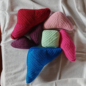May include: A selection of handmade crochet items in shades of red, pink, purple, blue, and green. The items are small, triangular shapes and a cube, all with a textured, ribbed pattern. They are arranged on a white fabric background, creating a colourful display.