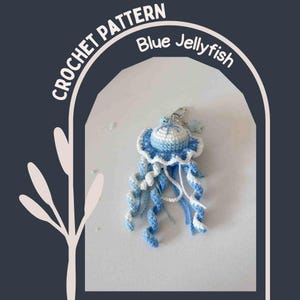 May include: A crocheted blue and white jellyfish keychain. The jellyfish has a round, domed head with a frilly edge and long, curly tentacles. The keychain is attached with a silver clasp. The text "CROCHET PATTERN Blue Jellyfish" is in the background.