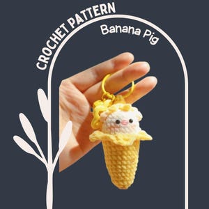 Op de afbeelding: Een gehaakte sleutelhanger in de vorm van een banaan met een varkensgezicht. De sleutelhanger is geel en wit, met de tekst "CROCHET PATTERN" en "Banana Pig". Het varken heeft een roze snuit en kleine zwarte ogen.
