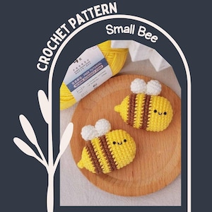 May include: Two small, crocheted bees with yellow and brown stripes and white wings rest on a wooden surface. The bees have smiling faces. A ball of yellow yarn and the text "CROCHET PATTERN Small Bee" are also visible.