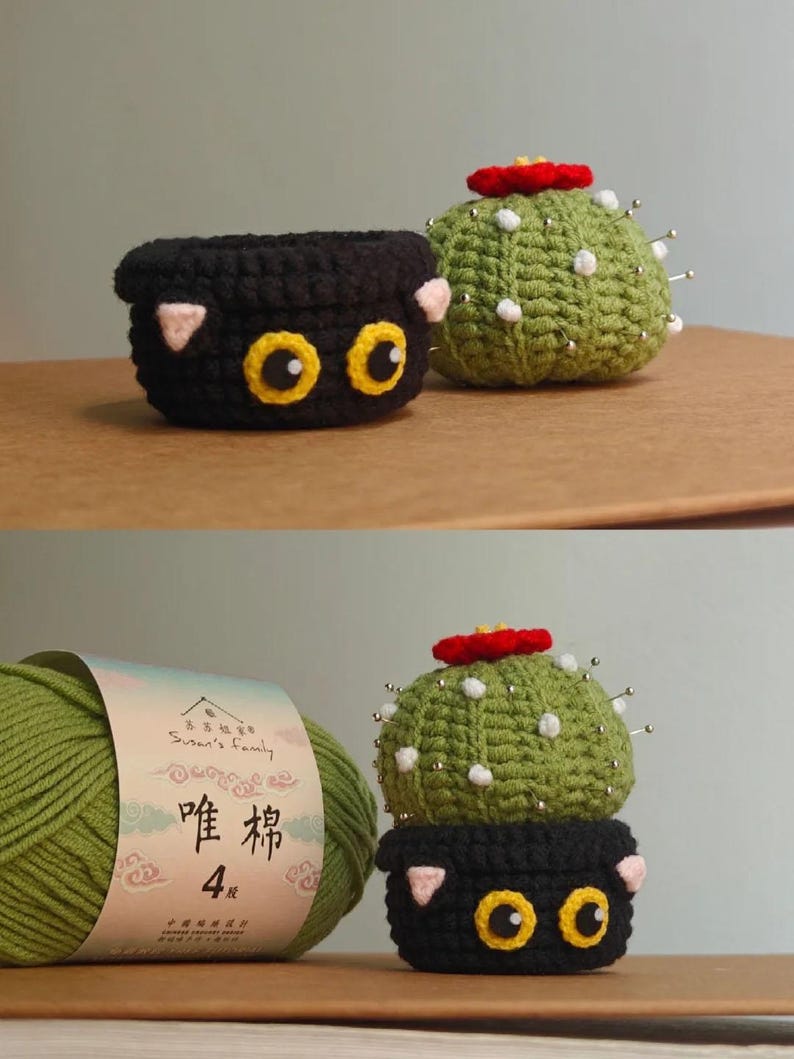May include: Crocheted cactus and cat-themed pincushions. The cactus is green with a red flower and white dots, and the cat pincushion is black with yellow eyes and pink ears. A skein of green yarn is also in the image.