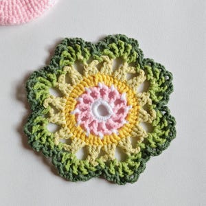 May include: A handmade, colorful crochet coaster in a floral design. The coaster features a central spiral of white and pink, surrounded by yellow, light green, and dark green crochet stitches. A small, pink crocheted item is visible in the upper left corner.