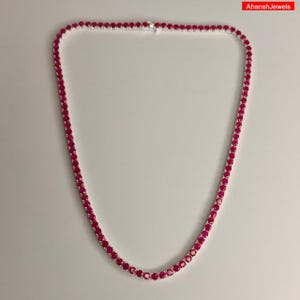 May include: A tennis necklace featuring a continuous row of round, vibrant pinkish-red stones set in a polished silver-toned metal chain. The necklace is arranged in a circular shape and includes a small metal clasp at the top center.