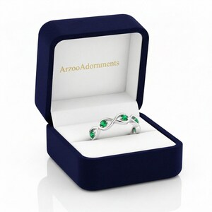 Op de afbeelding: Een zilveren ring met smaragdgroene edelstenen, gepresenteerd in een marineblauwe ringdoos. De ring heeft een uniek, vloeiend ontwerp. De doos is gevoerd met witte stof en heeft de tekst "ArzooAdornments" aan de binnenkant.