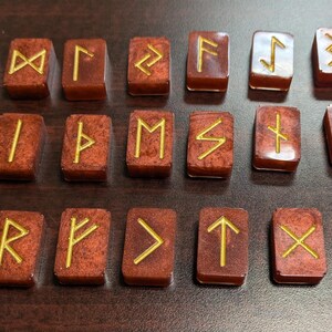 May include: A collection of 24 brown rune stones, each with a gold symbol. The rectangular stones are laid out on a dark surface. The symbols are likely used for divination or spiritual purposes.