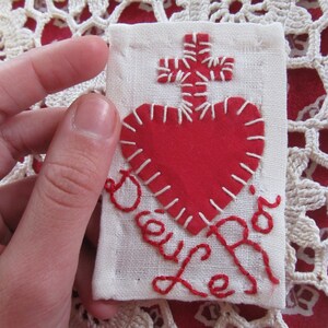May include: A white fabric square with red embroidery of a heart, cross, and the word "Joy" in cursive.