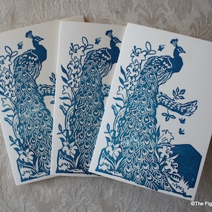 May include: Three blank greeting cards with a blue peacock design printed on them. The peacocks are surrounded by blue flowers and leaves. The cards are stacked on top of each other.