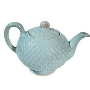 May include: A light blue ceramic teapot with a diamond pattern. The teapot has a curved handle, a spout, and a lid with a small knob. The teapot is a light blue color and is designed for brewing tea.