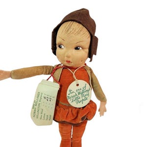 May include: A vintage Norah Wellings cloth doll wearing a brown felt hat, a tan jacket, and an orange velvet outfit. The doll features a round tag reading "I'm one of Norah Wellings' Little Pixie People" and a rectangular tag listing materials.