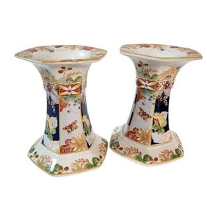 May include: A pair of hexagonal ceramic candle holders featuring a cream-colored base with intricate patterns of dark blue panels, orange and yellow floral motifs, and small brown butterflies, all accented with fine gold-colored trim details.