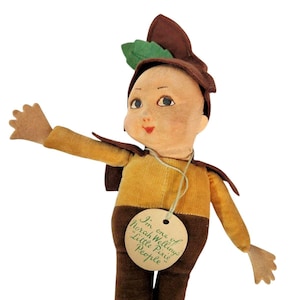 May include: A fabric doll with a tan face, painted features, and a brown hat adorned with a green leaf. The doll wears a yellow shirt and brown trousers, and features a round beige tag around its neck with the text: I'm one of Norah Wellings Little Pixie People.