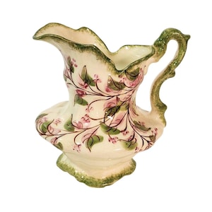 May include: Cream-colored ceramic pitcher with a green handle and rim. The pitcher is decorated with pink floral vines and green leaves. The base is also green. The pitcher has a unique shape with a spout and a wide opening.