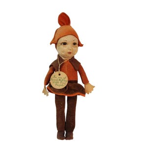 May include: A fabric doll with a pointed orange hat, wearing a brown and orange velvet outfit. A round beige tag hangs from the neck with the text: I'm one of Norah Wellings Little Pixie People. The doll has a painted face with dark eyes and hair.