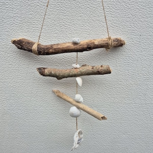 May include: A driftwood mobile, crafted with three horizontal wooden branches and twine. Small seashells and pebbles are attached, creating a natural, coastal aesthetic. The mobile hangs against a textured, light-coloured wall.
