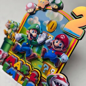 May include: A colorful, layered birthday cake topper featuring Mario and Luigi from the Super Mario Bros. video game. The design includes a number "2", coins, question mark blocks, and the names "Max & Emilio". The characters are in motion against a blue sky background.