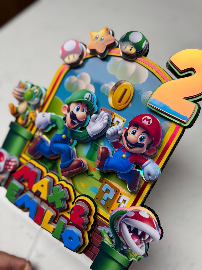 May include: A colorful, layered birthday cake topper featuring Mario and Luigi from the Super Mario Bros. video game. The design includes a number "2", coins, question mark blocks, and the names "Max & Emilio". The characters are in motion against a blue sky background.