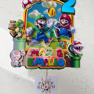 May include: A colorful cake topper featuring Mario and Luigi, the number 2, and the names "Max & Emilio." The design includes various Super Mario Bros. characters and elements, with a backdrop of a blue sky and green landscape.