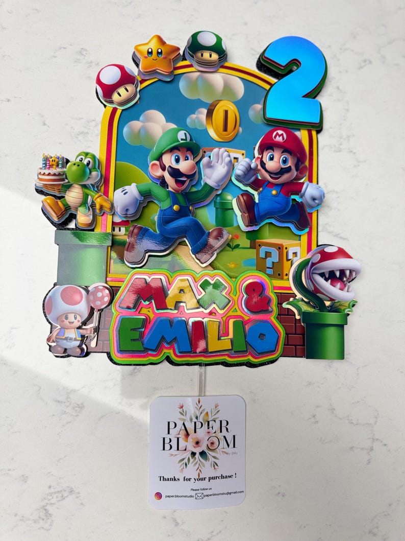 May include: A colorful cake topper featuring Mario and Luigi, the number 2, and the names "Max & Emilio." The design includes various Super Mario Bros. characters and elements, with a backdrop of a blue sky and green landscape.