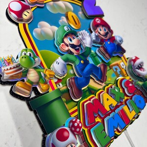 May include: A colorful cake topper featuring characters from the Super Mario Bros. video game. The design includes Mario, Luigi, Yoshi, and Toad, with a number 2 and the names "Max & Emilio". The background depicts a blue sky with clouds.