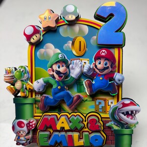 May include: A colorful cake topper featuring Mario and Luigi characters, a green Yoshi, and a Piranha Plant. The design includes a blue number 2, a gold coin, and the names "MAX & EMILIO". The background is a blue sky with clouds.