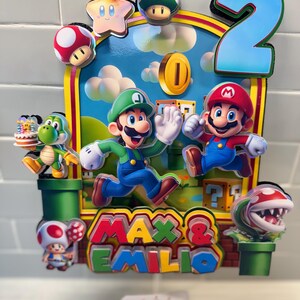 May include: A colorful birthday cake topper featuring Mario and Luigi characters, a star, mushrooms, and a gold coin. The number "2" is prominently displayed, along with the names "Max & Emilio". The background depicts a blue sky with clouds.