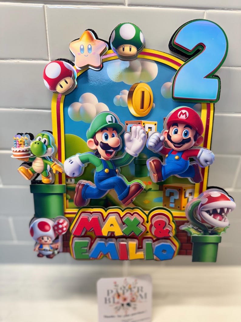 May include: A colorful birthday cake topper featuring Mario and Luigi characters, a star, mushrooms, and a gold coin. The number "2" is prominently displayed, along with the names "Max & Emilio". The background depicts a blue sky with clouds.