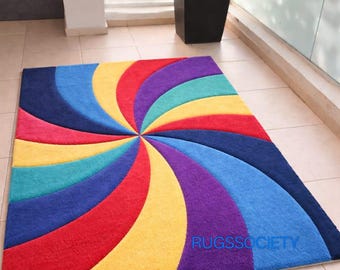 May include: A rectangular rug with a vibrant, swirling design in shades of red, yellow, blue, purple, and teal. The rug features a spiral pattern emanating from the center, with the text "RUGSSOCIETY" at the bottom.