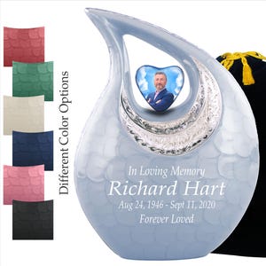 May include: A light blue memorial urn with a heart-shaped photo frame. The urn has a textured surface and silver accents. Text reads "In Loving Memory Richard Hart Aug 24, 1946 - Sept 11, 2020 Forever Loved". Different color options are shown.