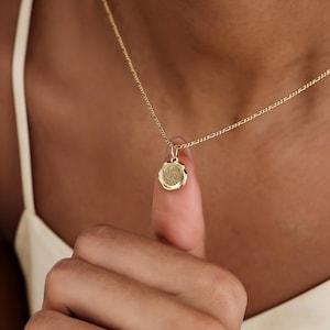 May include: A gold-colored necklace with a small, round pendant. The pendant has a textured surface and is suspended from a delicate chain. The necklace is displayed against a skin-toned background.