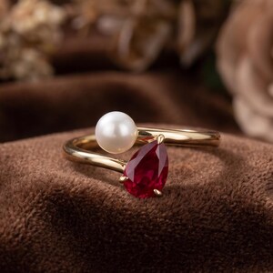May include: A gold bypass ring featuring a round white pearl on one end and a pear-shaped deep red gemstone on the other. The band is smooth and polished, creating an open design that showcases the contrast between the pearl and the red stone.