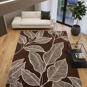 May include: A brown area rug with a large white leaf pattern. The rug is rectangular and covers a portion of a hardwood floor. A white sofa with a dark gray pillow sits in the background.