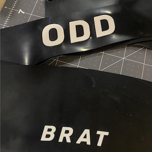 Könnte beinhalten: Schwarzes Gummi-Accessoire mit dem Wort "ODD" in weißen Blockbuchstaben. Ein weiterer Abschnitt des Zubehörs zeigt das Wort "BRAT" in weißen Blockbuchstaben. Das Zubehör hat einen Druckknopfverschluss.