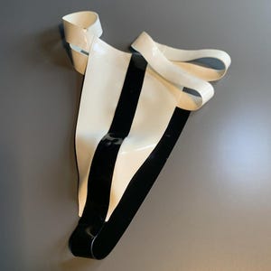 Könnte beinhalten: Ein Tanga aus glattem, glänzendem Material mit einem auffälligen Farbblock-Design. Das Kleidungsstück besteht aus einem breiten weißen Vorderteil mit einem vertikalen schwarzen Mittelstreifen, ergänzt durch weiße Seitenbänder und ein schwarzes Rückenteil.