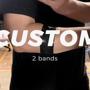 Puede incluir: Una banda negra para el brazo, usada en la parte superior, que presenta una superficie negra lisa y sólida con un pequeño emblema de cruz en tono plateado centrado. La banda es ancha y envuelve el brazo como una pieza única de material oscuro.