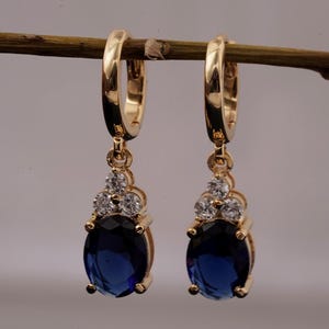 Peut inclure: Une paire de boucles d'oreilles créoles dorées avec un design pendant. Chaque boucle d'oreille présente une grosse pierre précieuse ovale bleu foncé, ornée de petites pierres claires. Les boucles d'oreilles sont suspendues à une créole dorée.