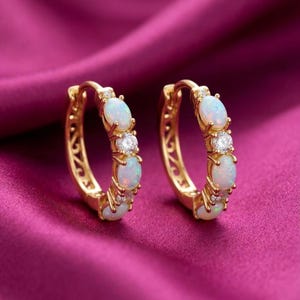 May include: Gold hoop earrings with opal and diamond detailing. Each earring has three oval opals and two small diamonds, set in a gold frame. The earrings are presented on a deep pink satin background.